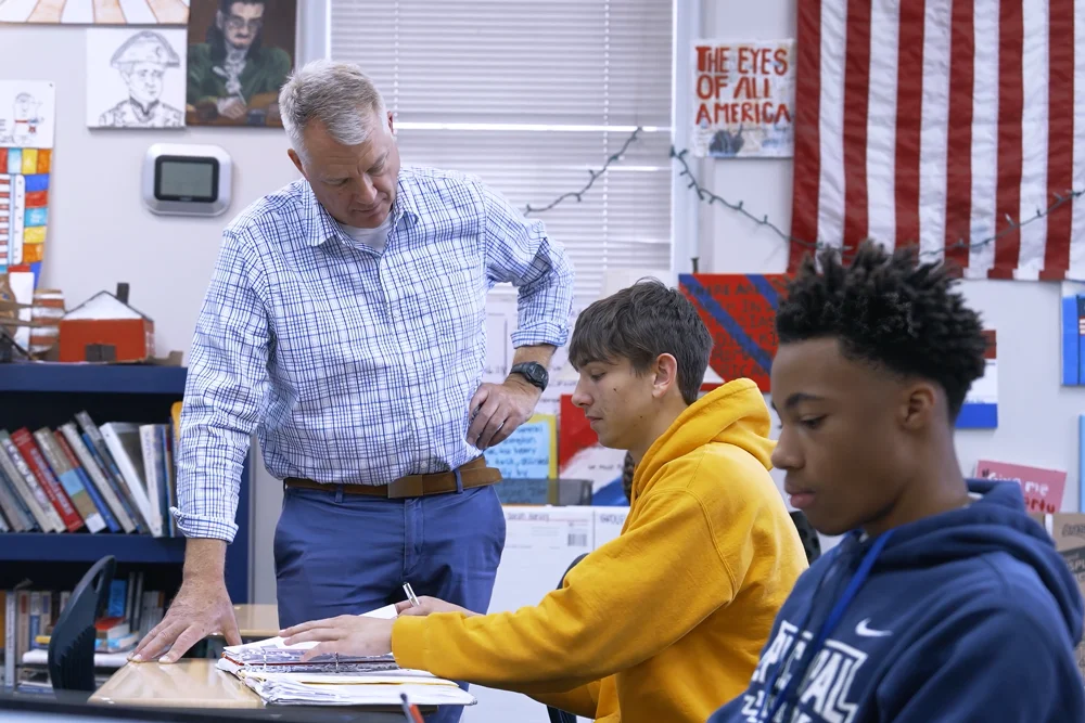 Teacher leaning over a student's desk to help while another student works; classroom with bookshelves and an American flag in the background.