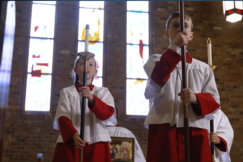 Two altar servers in white surplices and red robes holding tall processional candles in front of stained-glass windows