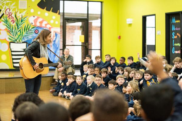 woman with guitar singing to a group of students in room with yellow walls