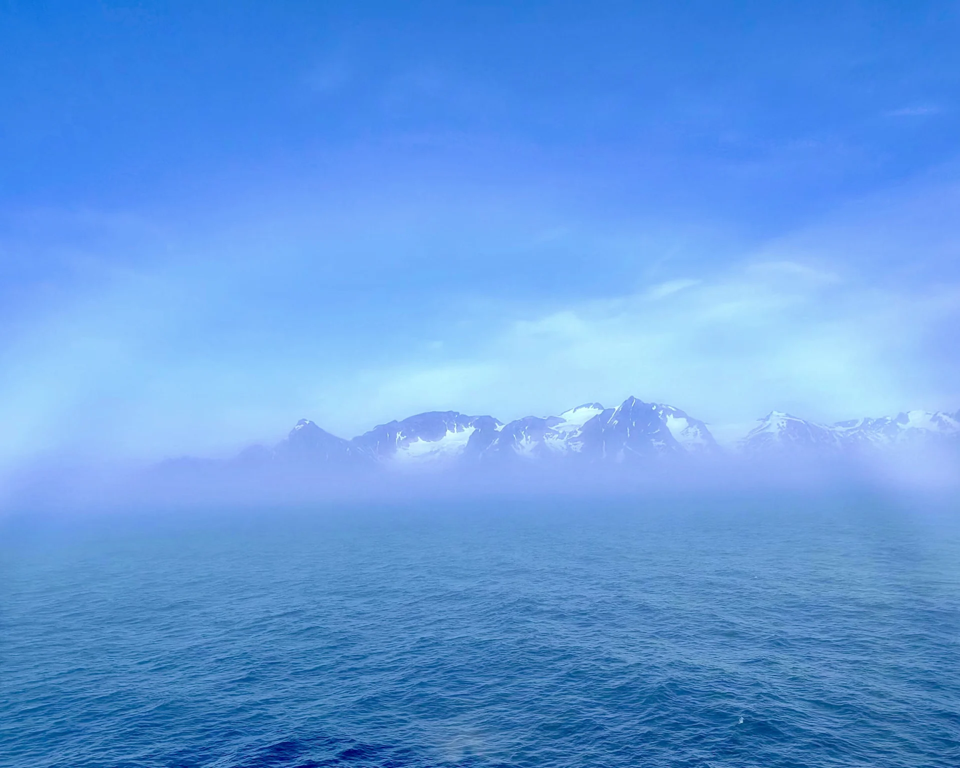 Calm open sea with a misty horizon and distant snow‑capped mountains beneath a clear blue sky
