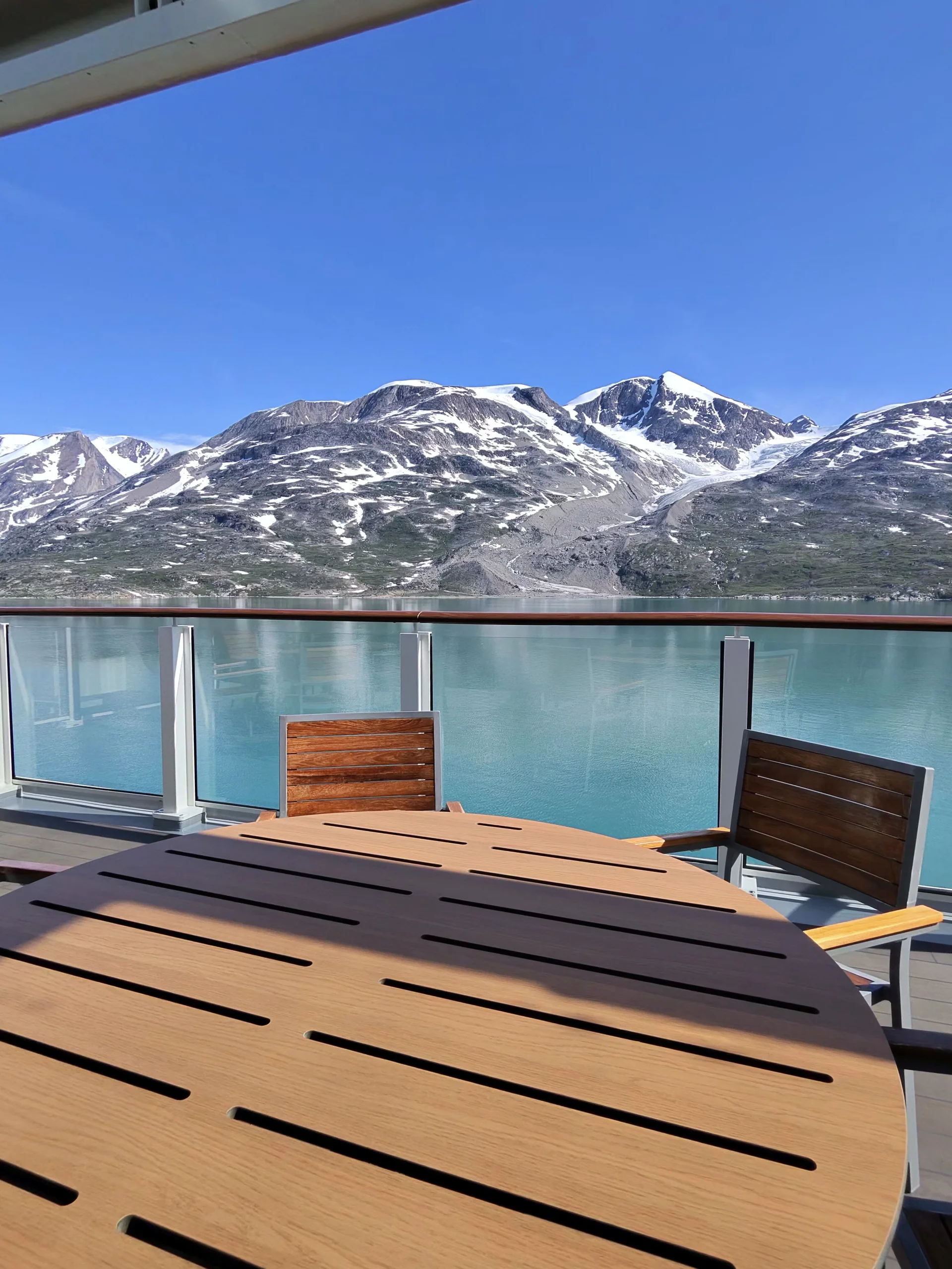 Wooden table and chairs on a balcony deck overlooking a turquoise fjord and snow‑capped mountains under a clear blue sky.