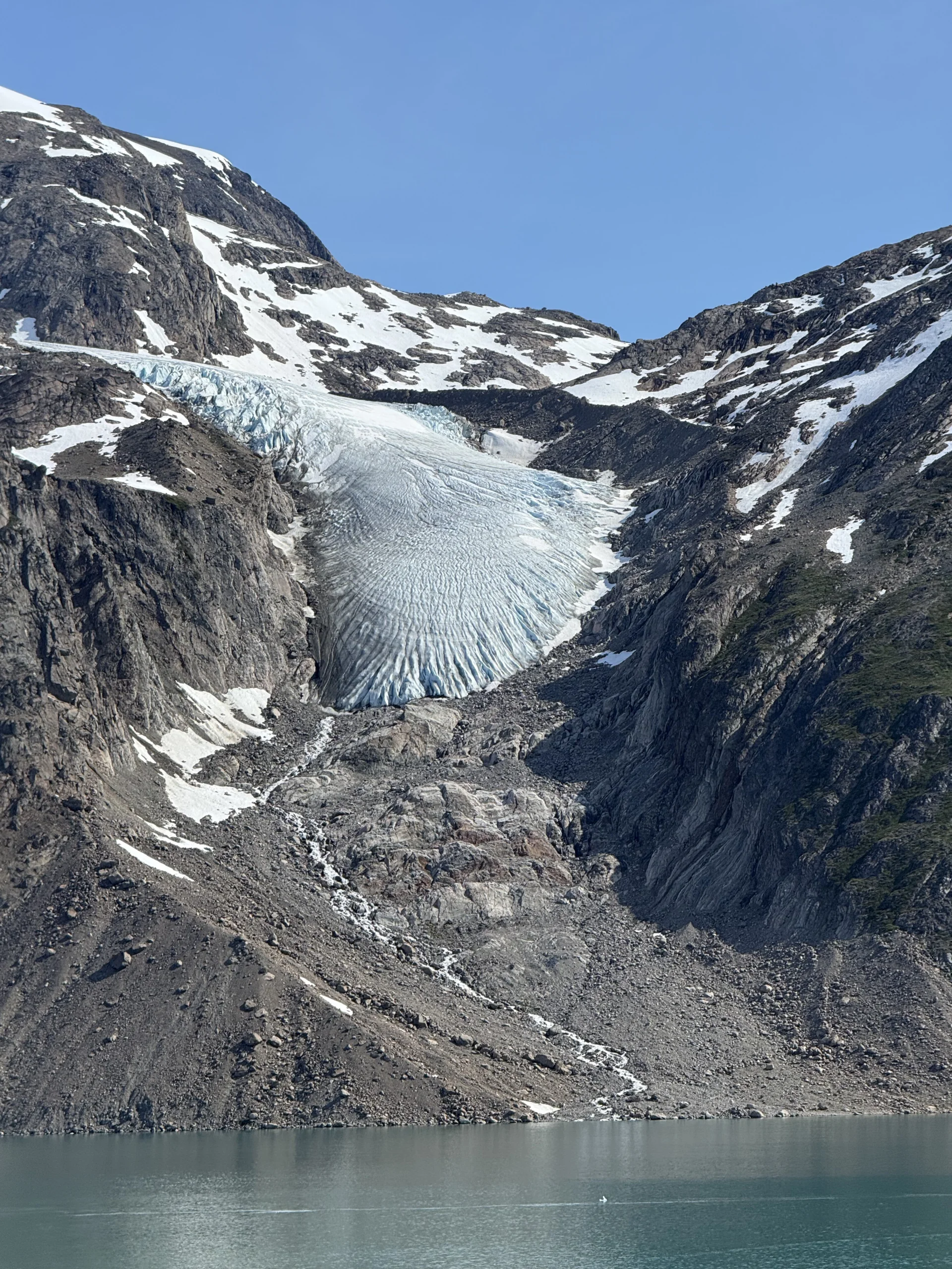 Glacier snout and surrounding rock slopes above an alpine lake Crevassed glacier tongue descending between rocky, snow‑patched mountains toward a turquoise glacial lake