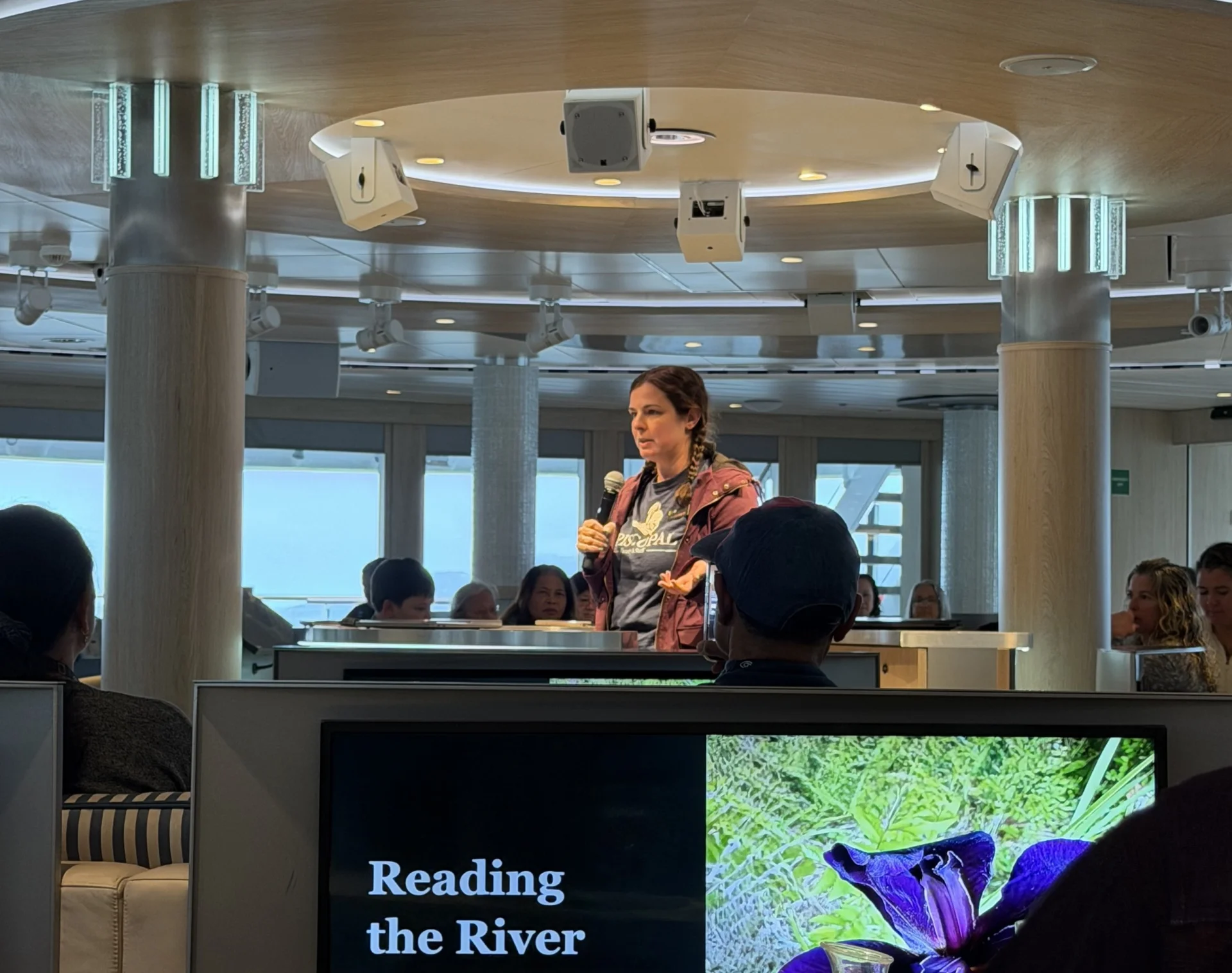 Woman holding a microphone speaking to seated passengers in a wood‑paneled lounge; a large screen in the foreground shows the title “Reading the River.”