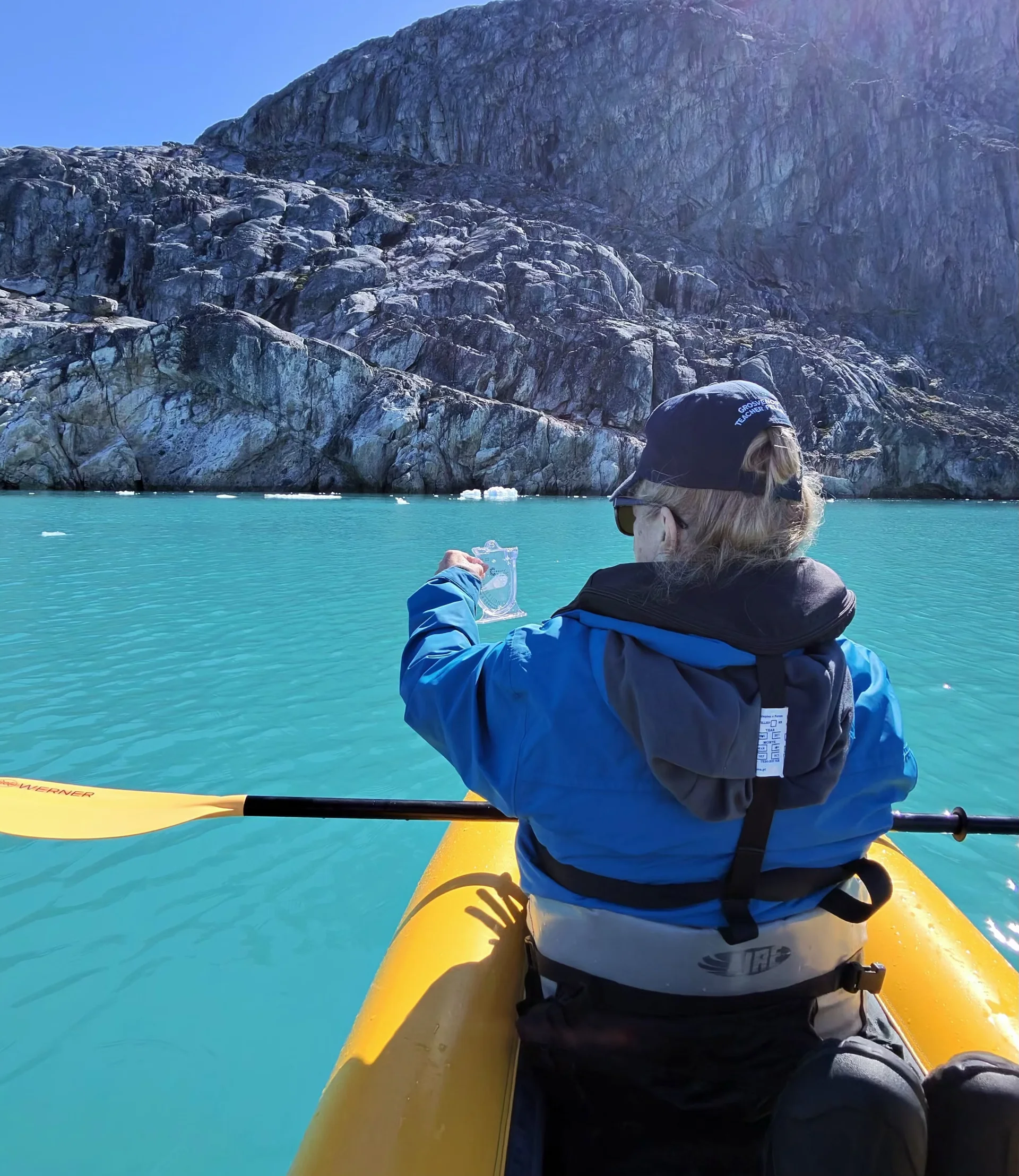 Person wearing a cap and blue jacket in a yellow kayak on bright turquoise glacial water, holding a small clear fragment of ice toward a steep rocky cliff