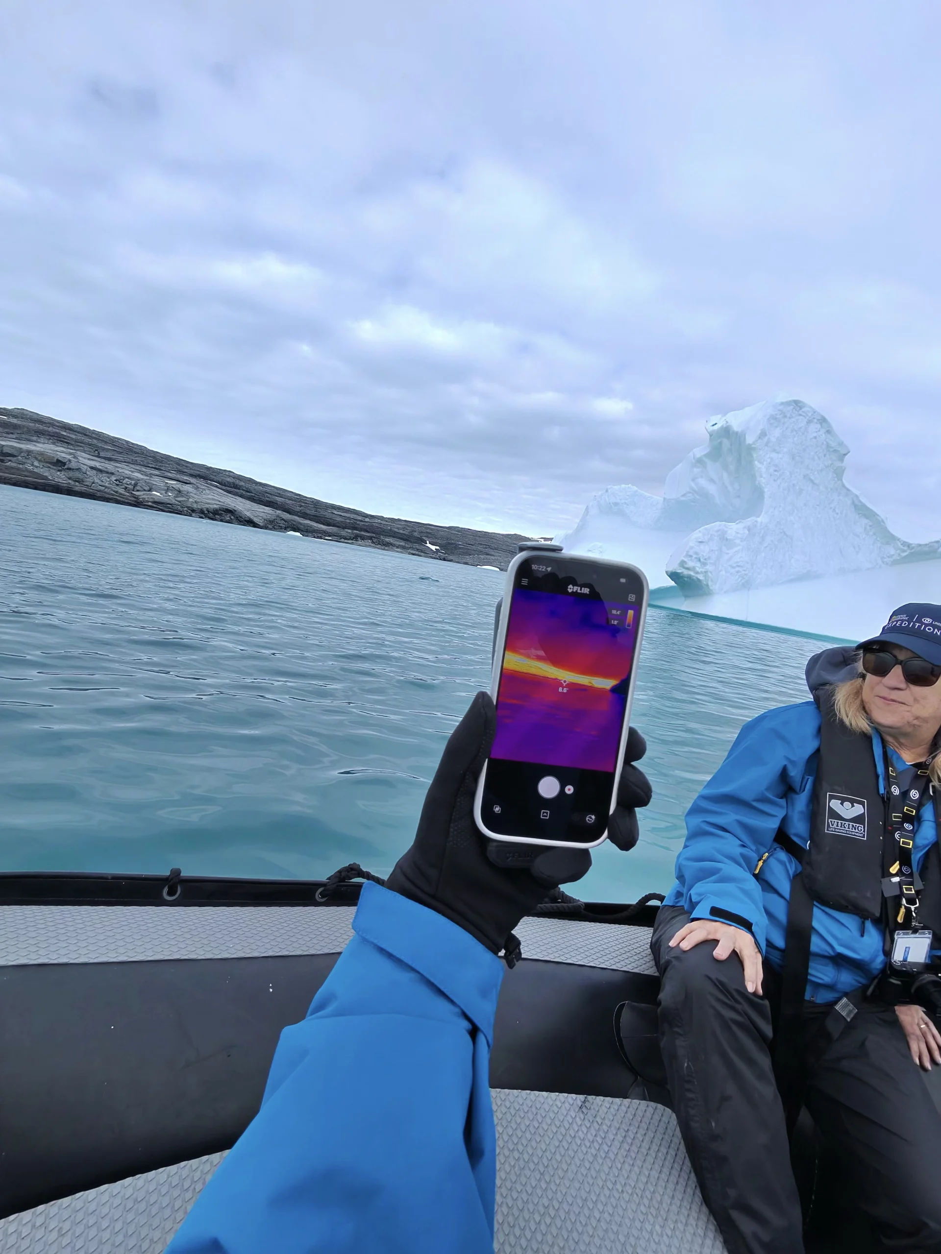 Person in a small inflatable boat holding a thermal-imaging smartphone toward a large iceberg and rocky shoreline; a companion in a blue jacket and life vest sits to the right.