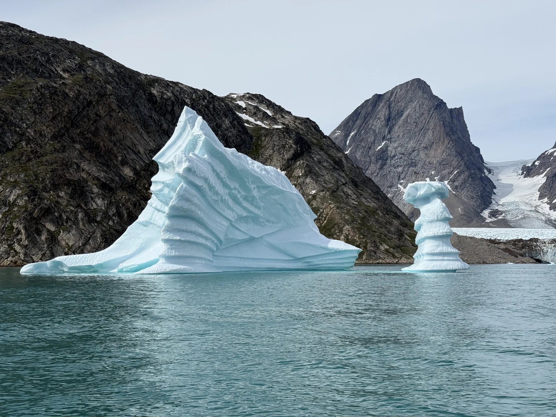 Panoramic photo emphasizing iceberg shapes against a glacial coastline Two sculpted blue-white icebergs floating in calm teal water before steep rocky mountains and a glacier