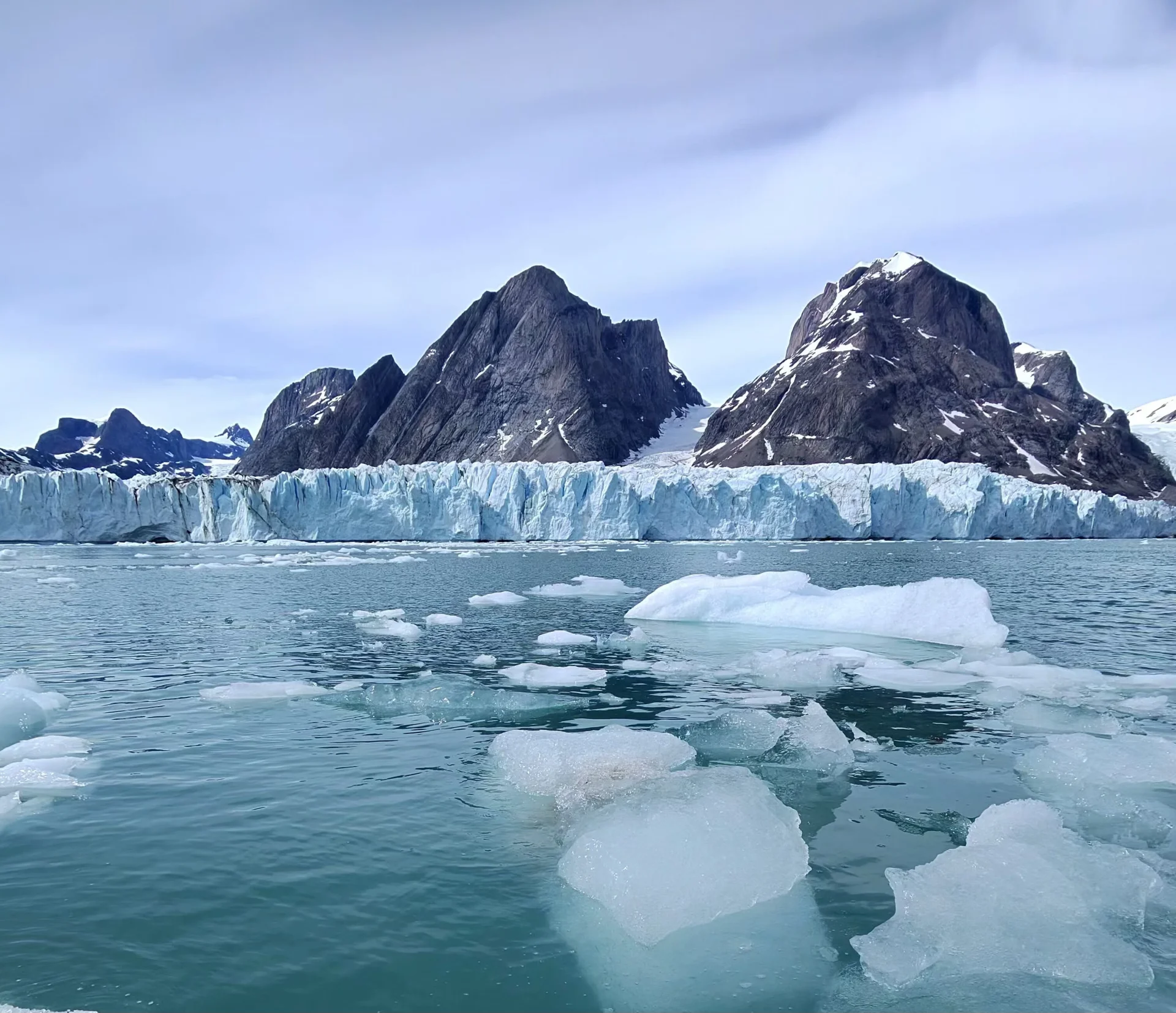Blue glacier front with floating ice chunks in teal water and dark, snow-dusted mountains beneath a pale sky.