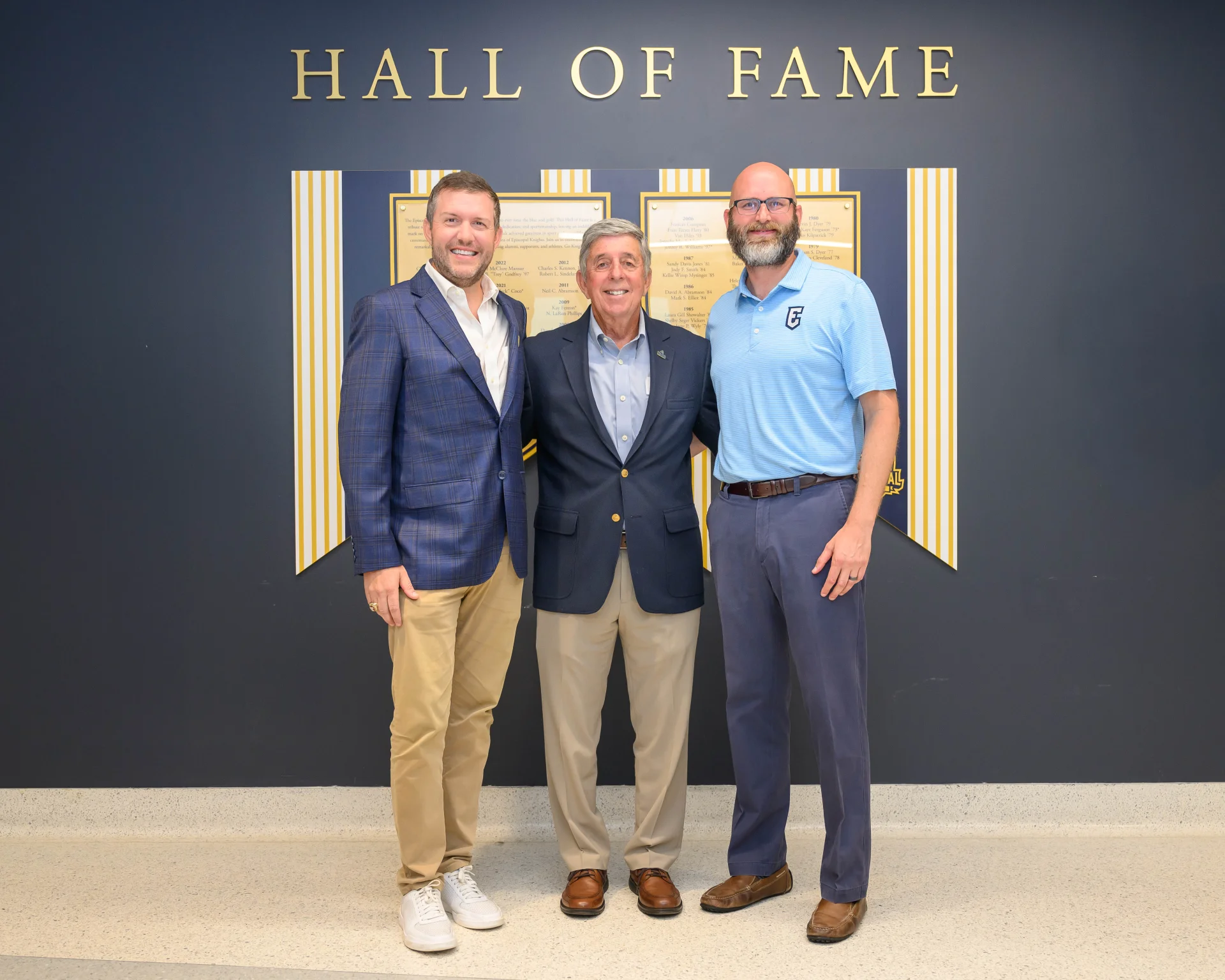 Three adult men smiling and standing together in front of a 'HALL OF FAME' wall that displays framed plaques