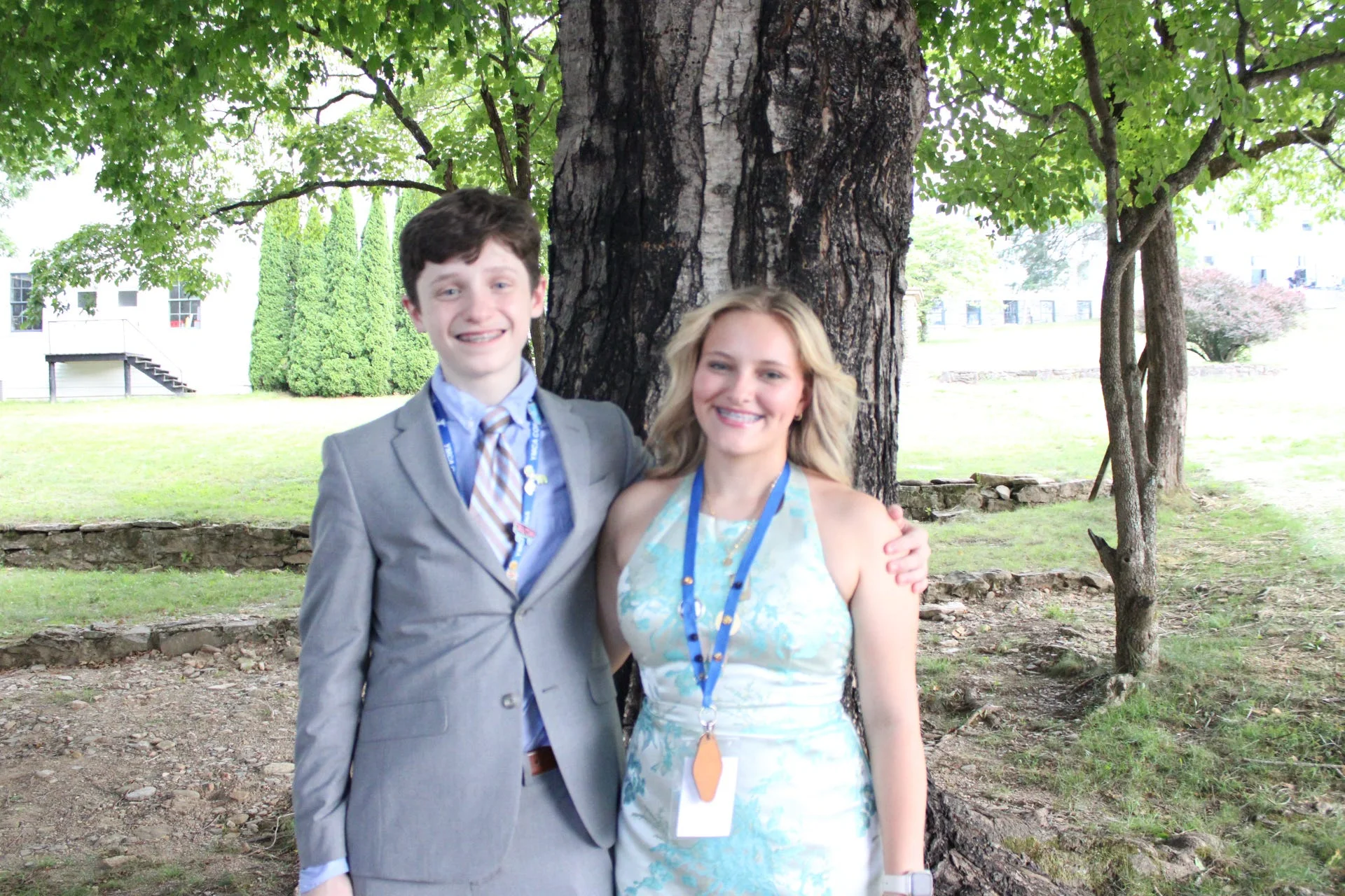 Young man in a gray suit and young woman in a light blue dress wearing lanyards, smiling with arms around each other in front of a large tree