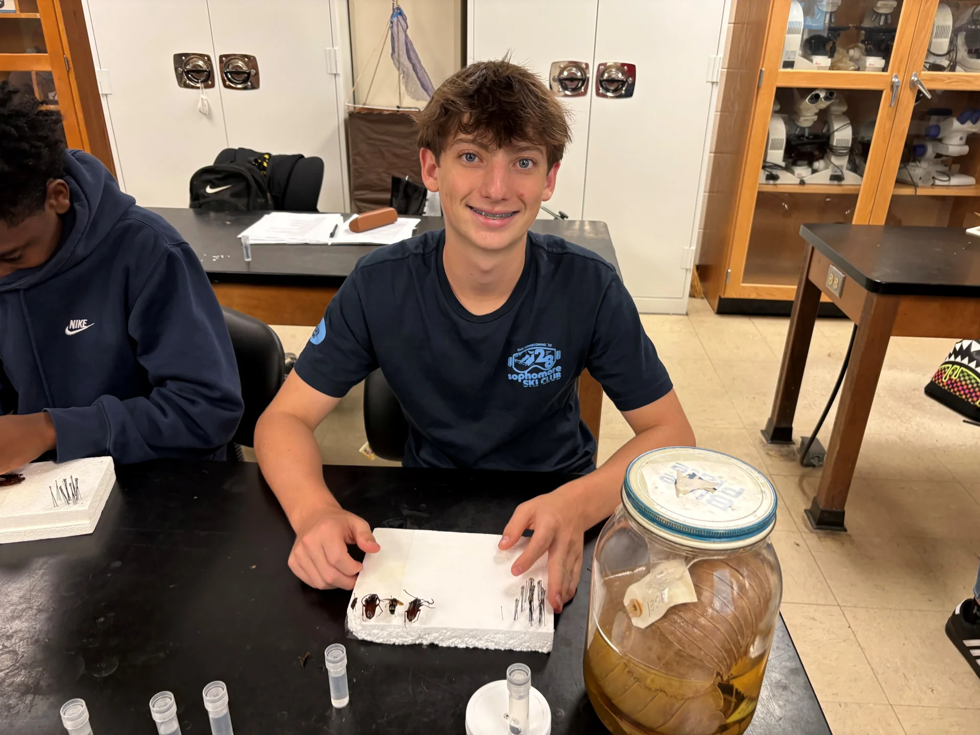 Smiling student seated at a lab table arranging pinned beetle specimens on white foam; large jar with a preserved specimen at right and another student partly visible at left