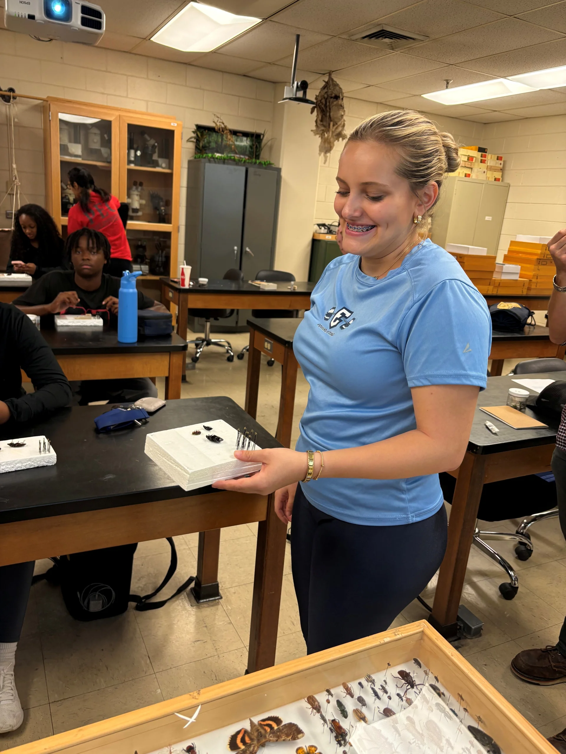 Student presenting pinned insect specimens in a classroom entomology lab Smiling student holding a foam block with pinned insect specimens while standing at a lab table; a display tray of pinned insects and classmates are visible in the background.