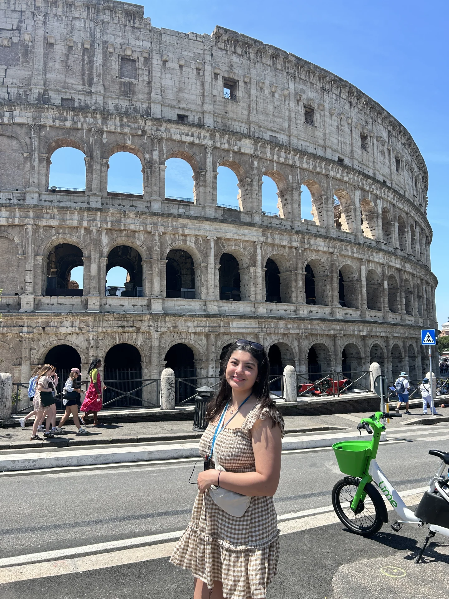 Young woman smiling near a crosswalk with the Colosseum's arched facade behind her and a green rental scooter at right.