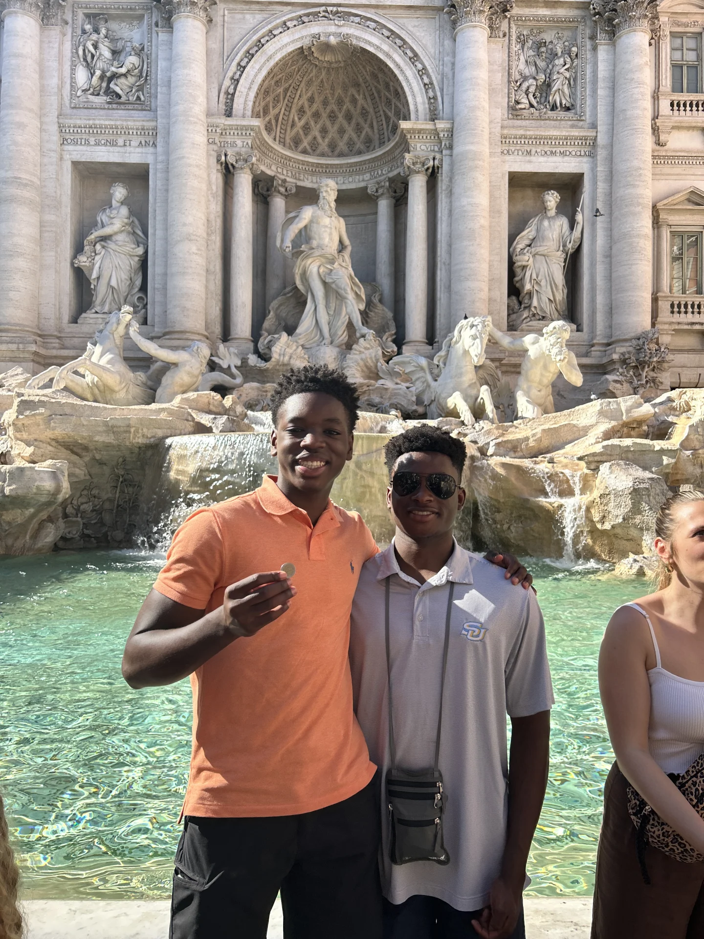 Trevi Fountain (Rome): two young men posing, one holding a coin Two young men smiling and posing in front of an ornate fountain; one holds a coin toward the camera with carved statues and flowing water behind them.