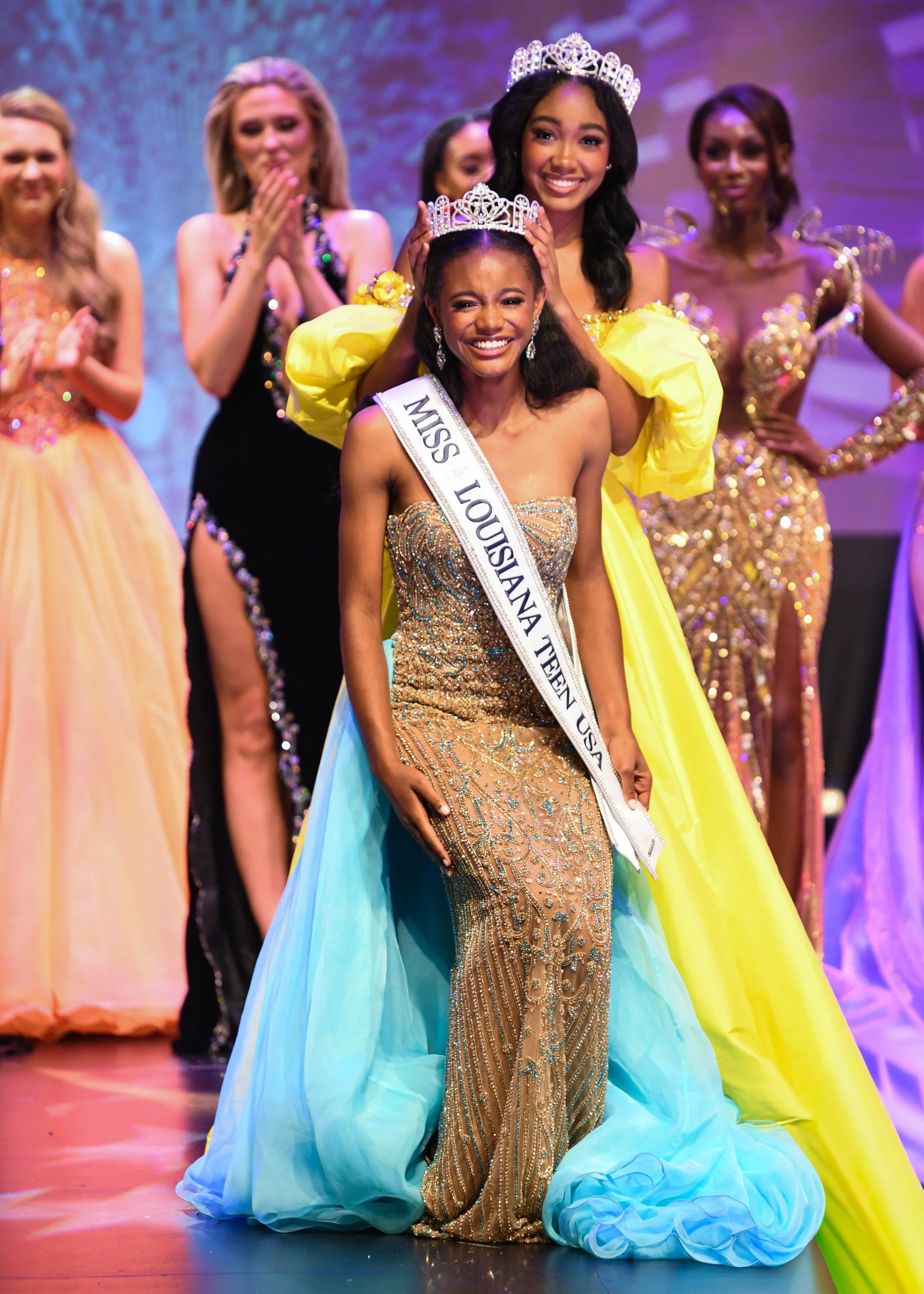 Smiling pageant winner being crowned onstage, wearing a beaded gold gown with a blue train and a sash reading “Miss Louisiana Teen USA” while other contestants applaud.