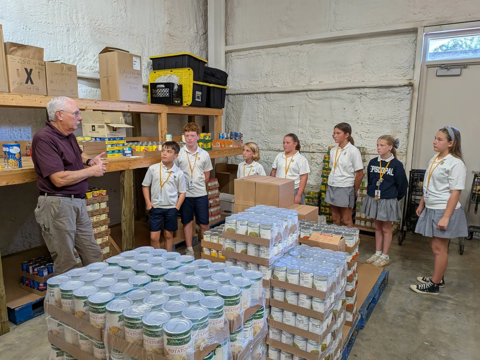School group listening to a volunteer’s instructions at a food pantry. Older volunteer speaking to a line of school students beside shelves and pallets of canned goods in a food pantry.
