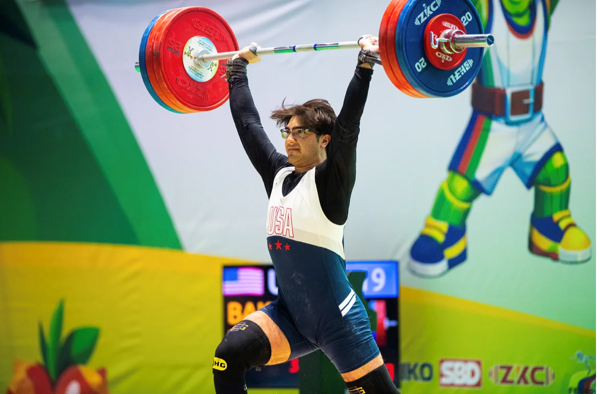 Competition stage with scoreboard and colorful mascot backdrop Athlete wearing a USA singlet and glasses holding a heavily loaded barbell overhead in a split-stance lift