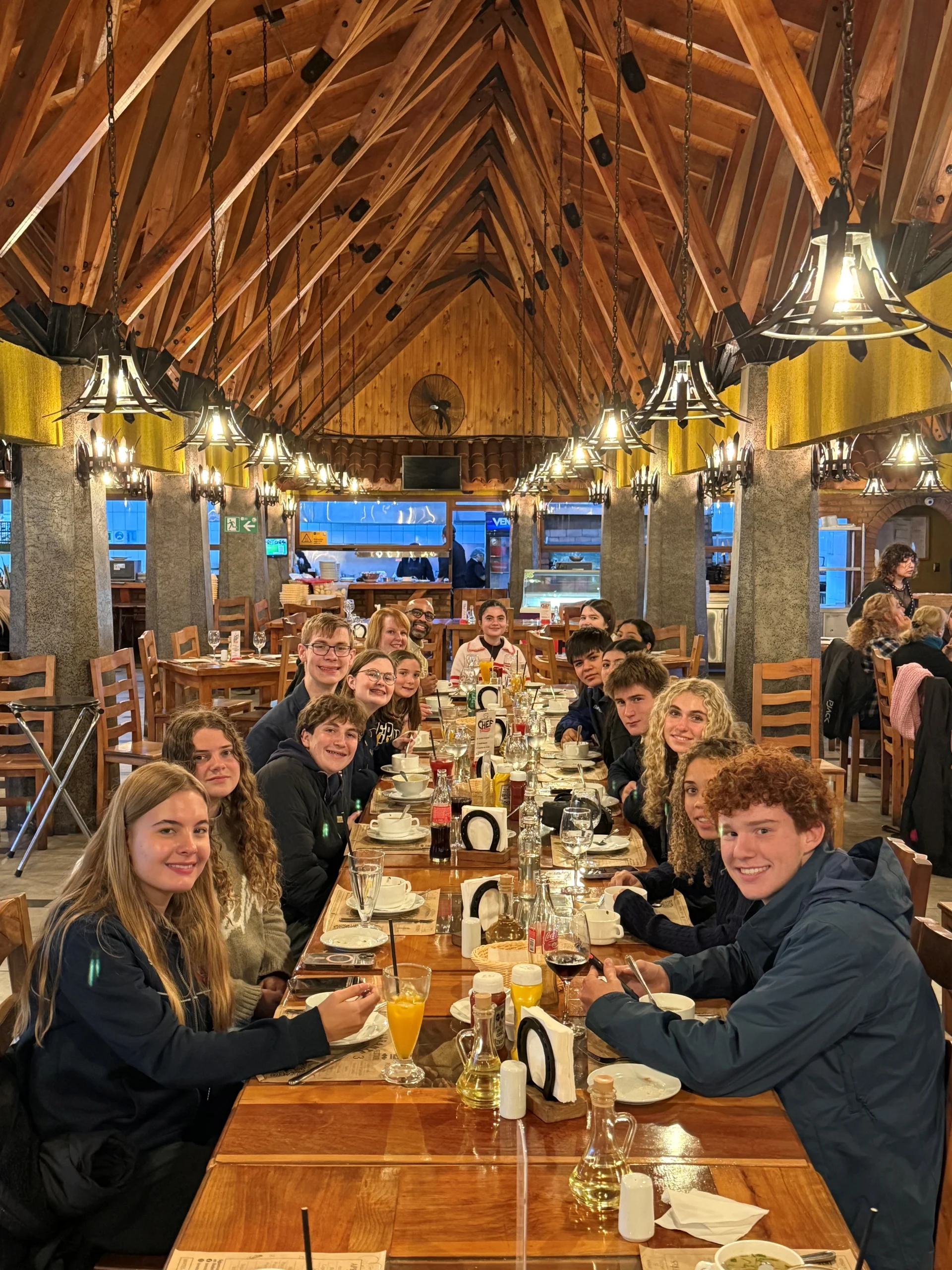 Group of smiling teenagers and adults seated around a long wooden dining table in a rustic restaurant with a vaulted wooden ceiling and hanging lanterns.