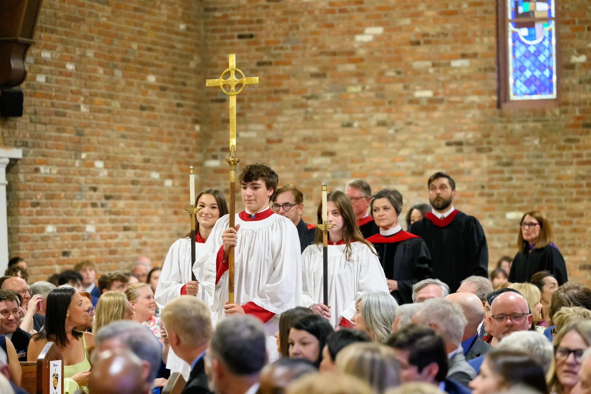 Acolytes leading a church procession Three youth acolytes in white robes with red collars lead a church procession carrying a brass cross and two candles, congregation and clergy behind them.