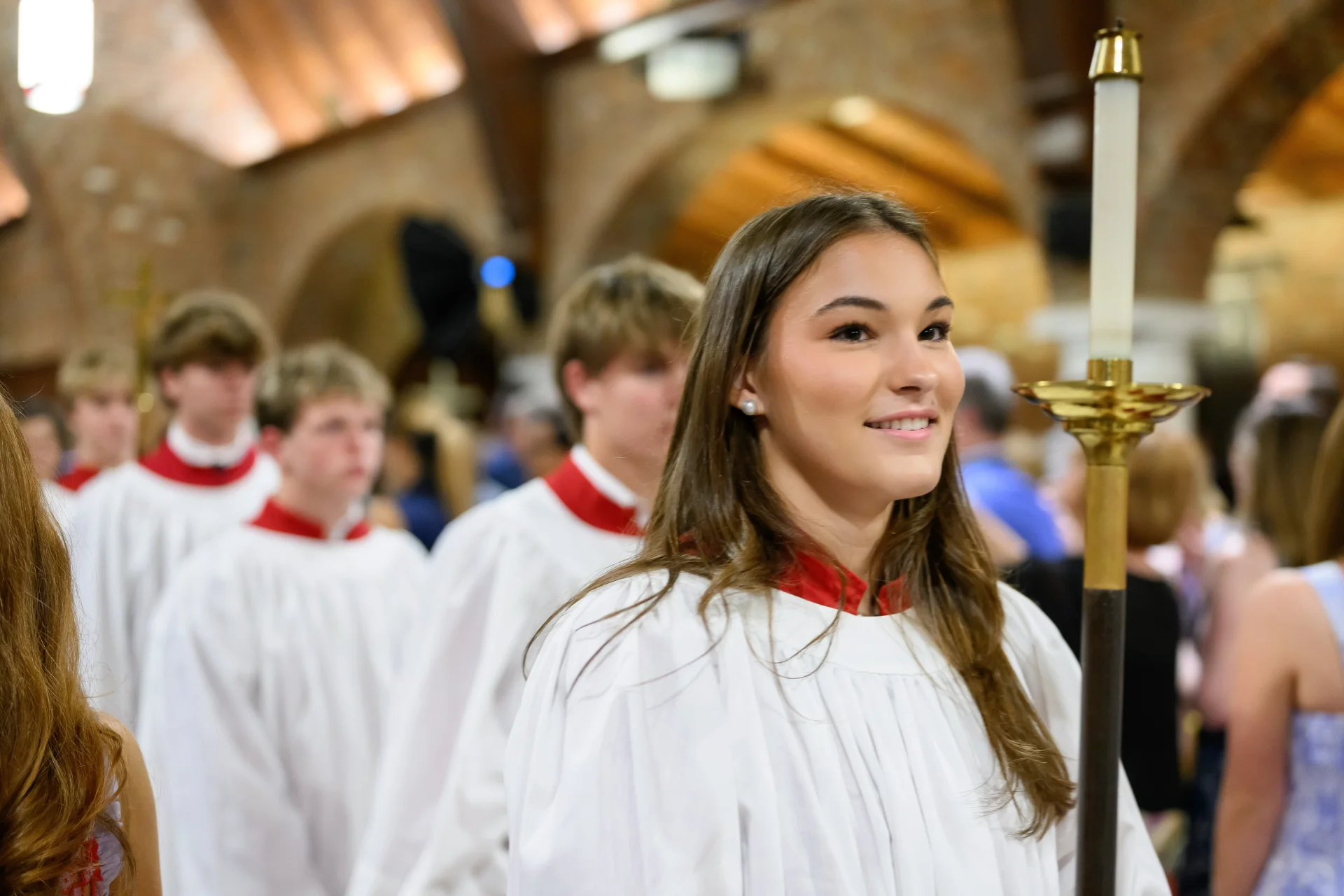 Smiling young acolyte in a white robe with a red collar holding a tall processional candle, with fellow acolytes following in a church interior