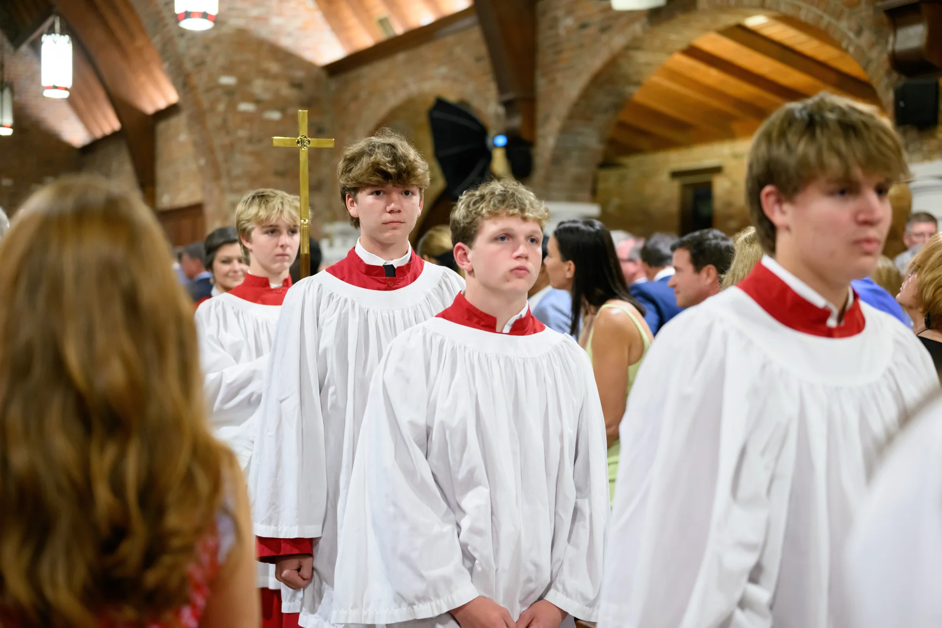 Three acolytes in white robes with red collars process down a church aisle; one holds a brass processional cross while worshippers stand along the sides.